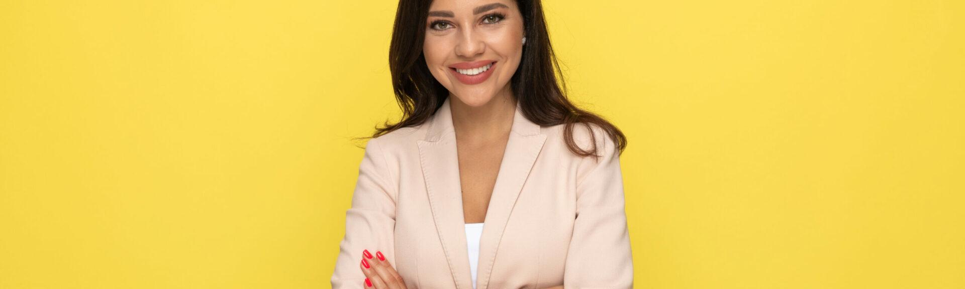 happy young businesswoman in pink suit crossing arms and smilling on yellow background